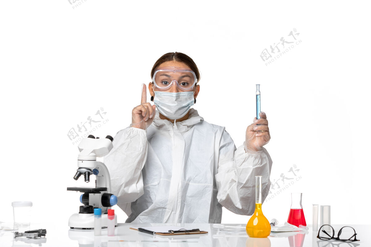 创意正面图：女医生穿着防护服 戴着口罩 在白色桌子上拿着装有病毒冠状病毒溶液的瓶子视图正面面罩