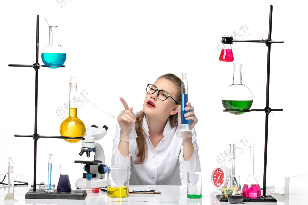 健康前视图穿着医疗服的女化学家拿着长烧瓶 白色背景上有蓝色溶液化学大流行健康病毒医生套装前面