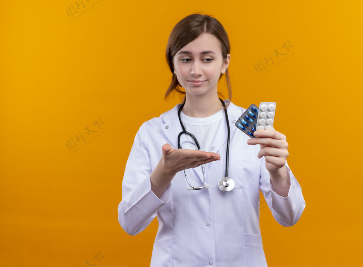 年轻自信的年轻女医生 穿着医用长袍 手持听诊器 手持药品 用手指着他们 站在隔离的橙色空间和复印空间里医学听诊器长袍