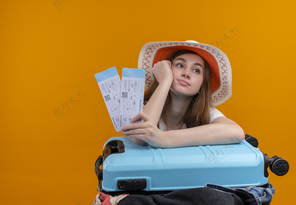 女孩无聊的年轻旅行家女孩戴着帽子拿着机票把手放在脸颊上 胳膊放在手提箱上孤立的橙色空间与复制空间空间持有手臂