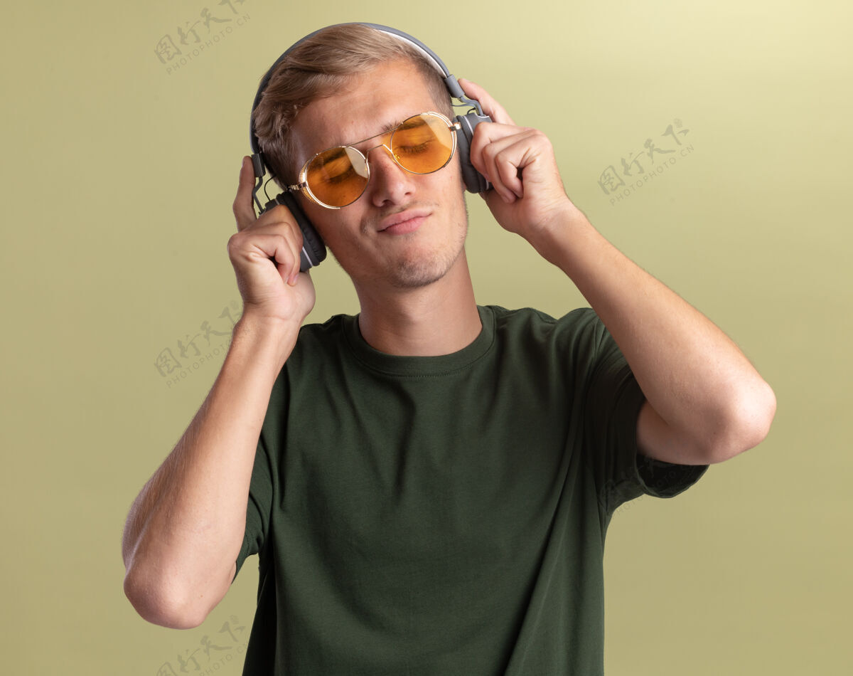 耳机喜欢闭着眼睛的年轻帅哥穿着绿色衬衫 戴着眼镜和耳机 隔离在橄榄绿的墙上脸橄榄人