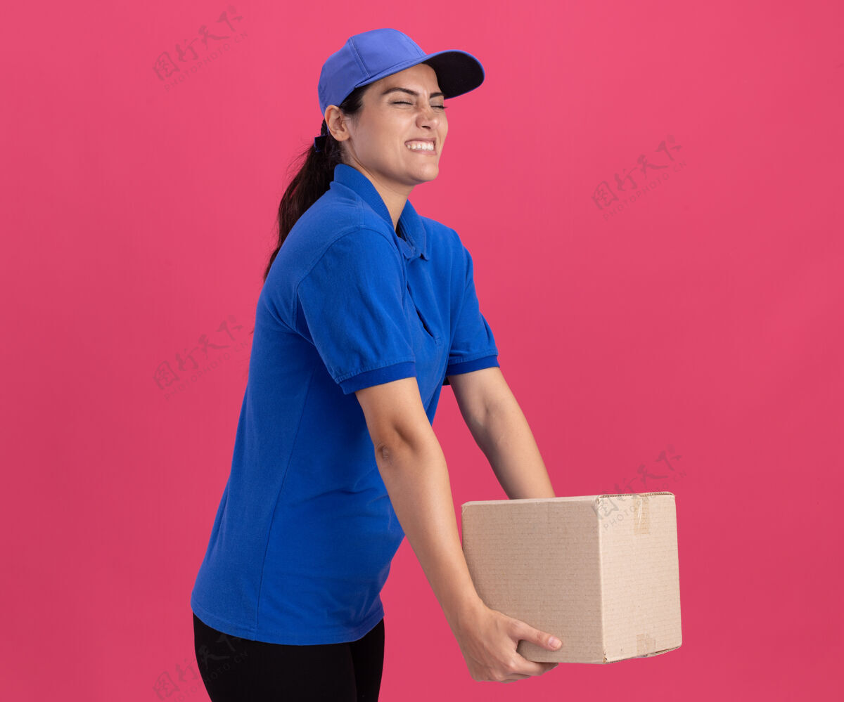 紧张紧闭着眼睛紧张的年轻送货女孩穿着制服 粉色墙上隔离着帽子盒表情持有姿势