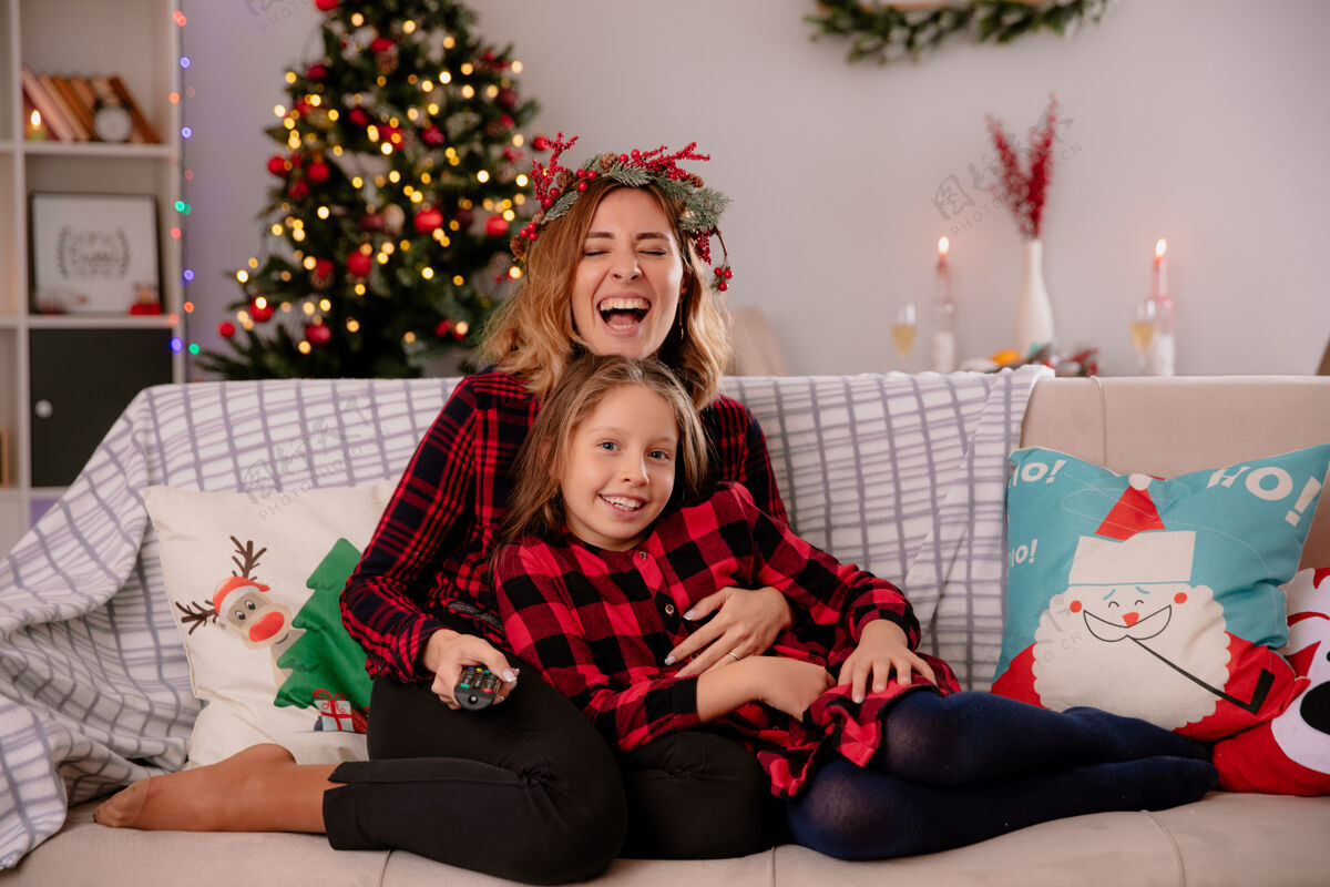 享受带着冬青花环的欢笑妈妈拿着电视遥控器 看着相机 女儿坐在沙发上 在家里享受圣诞节时光圣诞快乐举行花圈