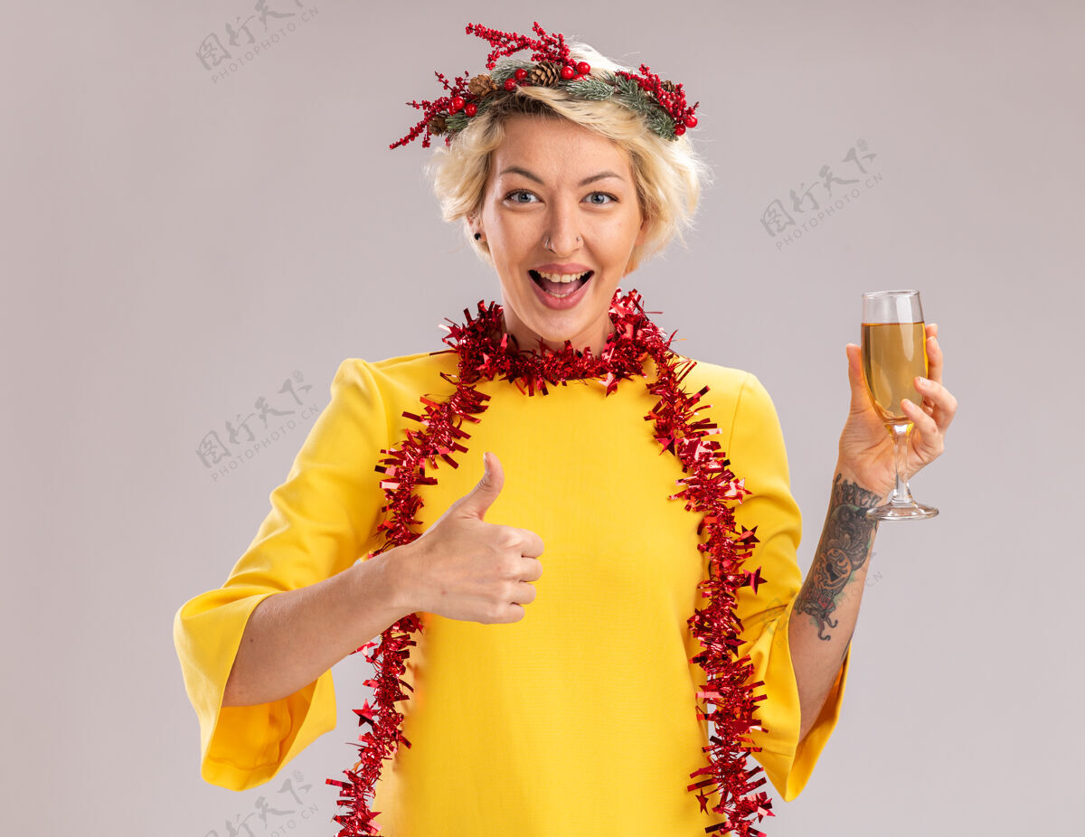 圣诞快乐快乐的金发女郎戴着圣诞花环 脖子上戴着金箔花环 手里拿着一杯香槟 看着镜头 在白色背景上孤立地竖起大拇指欢乐头年轻