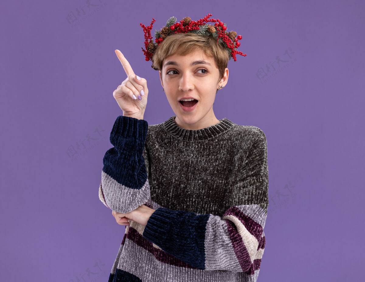 圣诞快乐令人印象深刻的年轻漂亮女孩戴着圣诞花环看着一边指着孤立的紫色背景复制头指向
