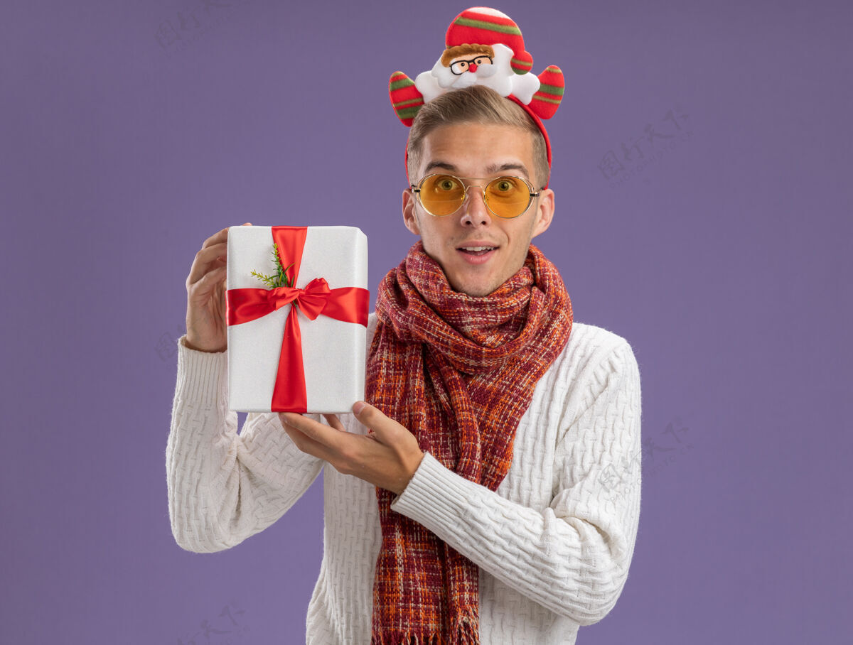 复制印象深刻的年轻帅哥戴着圣诞老人的头带和围巾拿着礼包看着隔离在紫色背景上的相机头带圣诞快乐年轻