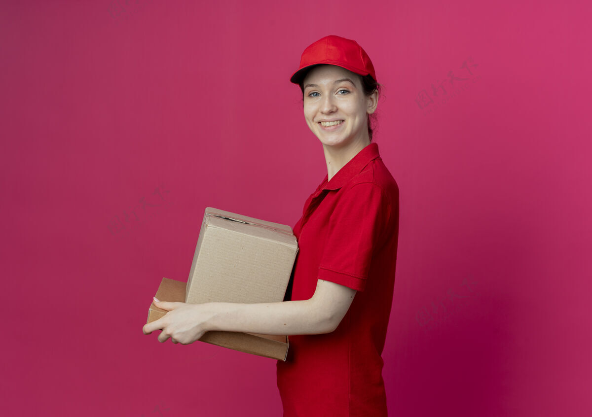 盒子微笑着的年轻漂亮的送货女孩穿着红色制服 戴着帽子站在侧视图中 拿着纸箱和披萨包 在深红色背景上与复印空间隔离开来年轻红色送货