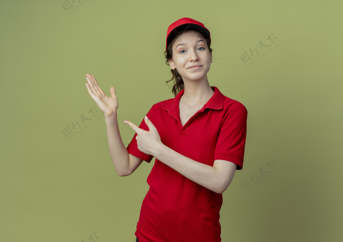 手高兴的年轻漂亮的送货女孩在红色制服和帽子显示空手 并在橄榄绿的背景与复制空间孤立的一面指着女孩指向橄榄