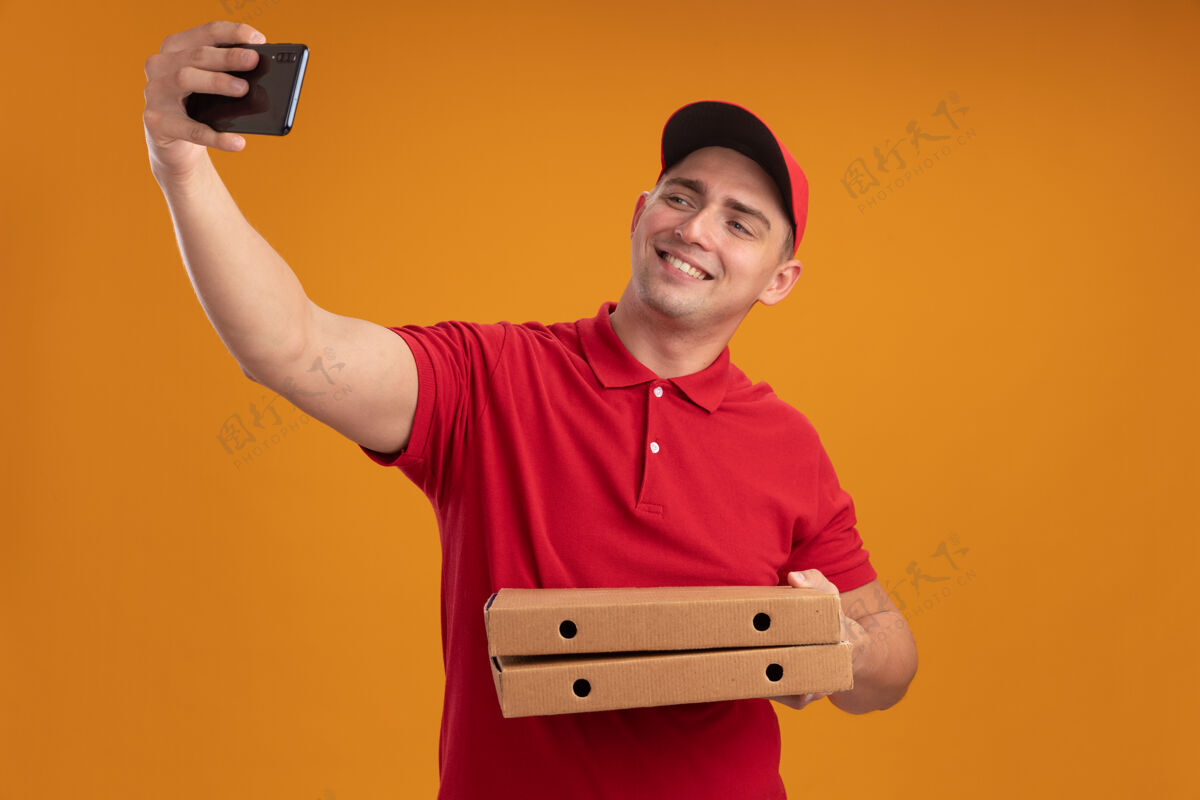 微笑微笑的年轻送货员穿着制服 戴着帽子 拿着披萨盒 在橙色的墙上自拍帽子感觉抱着