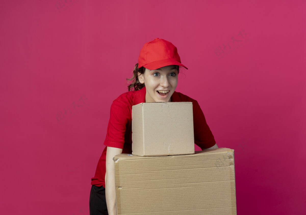 制服令人印象深刻的年轻漂亮的送货女孩在红色制服和帽子看直举行纸箱孤立的深红色背景与复制空间印象年轻直