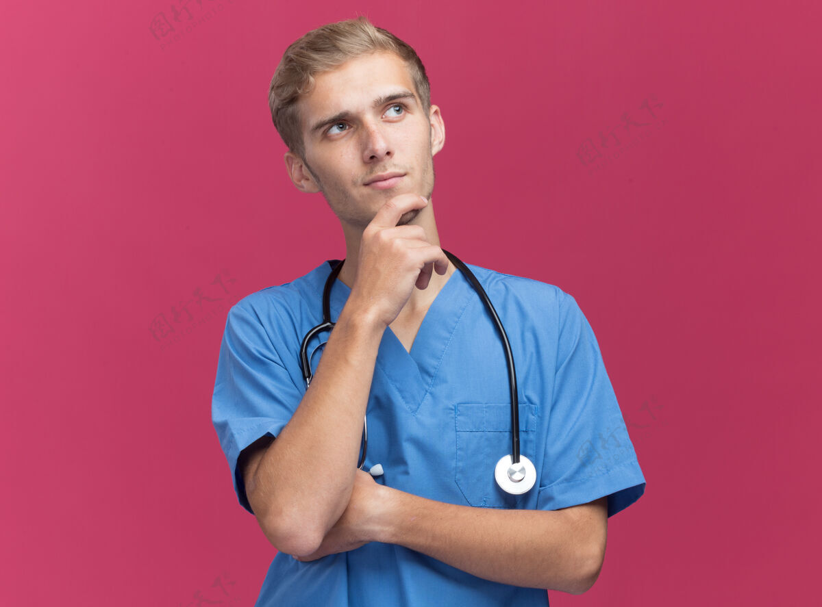 制服令人印象深刻的看一边年轻的男医生穿着医生制服听诊器举行下巴隔离在粉红色的墙上脸印象下巴
