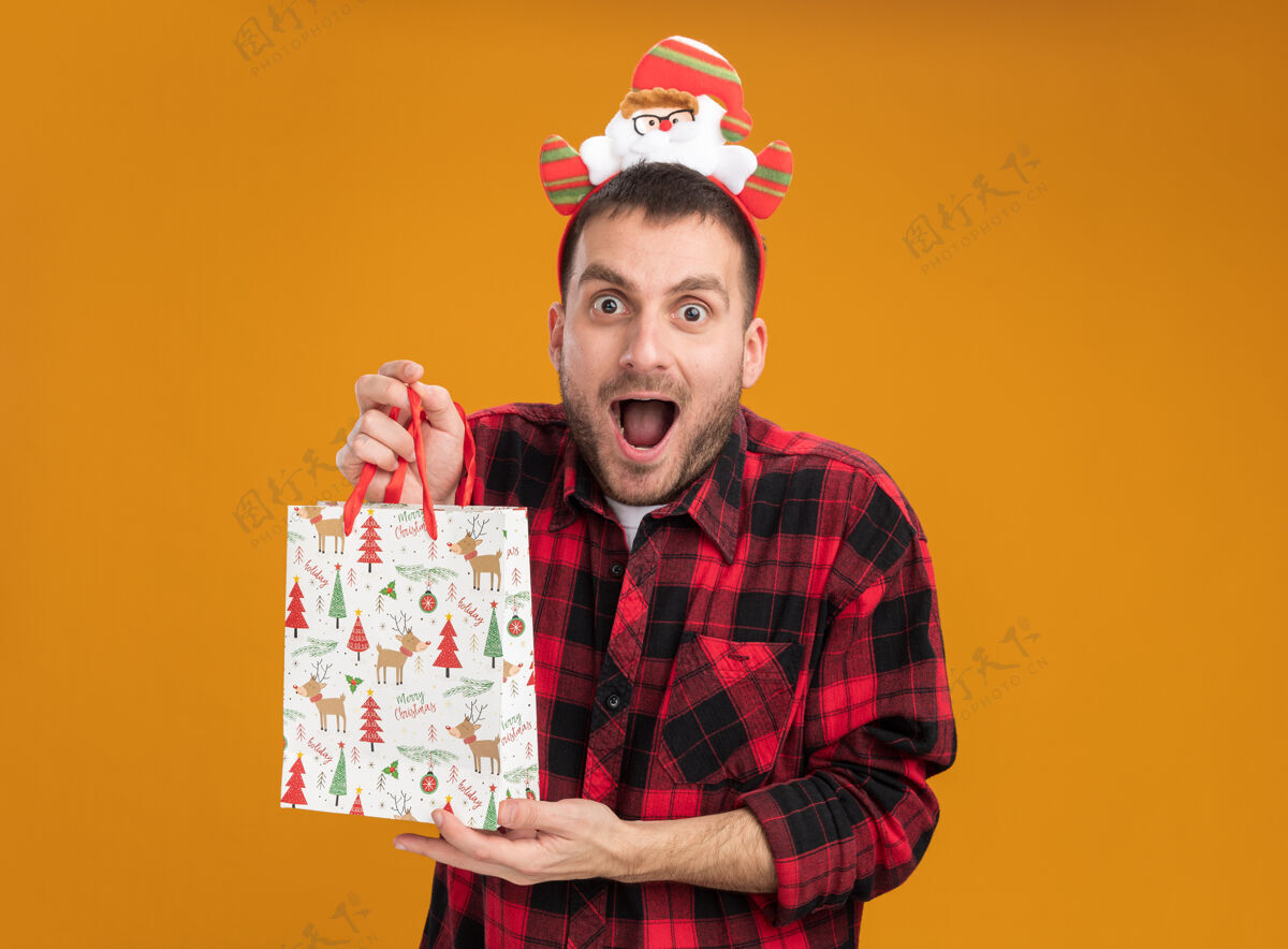 包令人印象深刻的年轻白种人戴着圣诞老人的头带 拿着圣诞礼品袋 看着橙色背景上孤立的摄像机空间年轻礼物