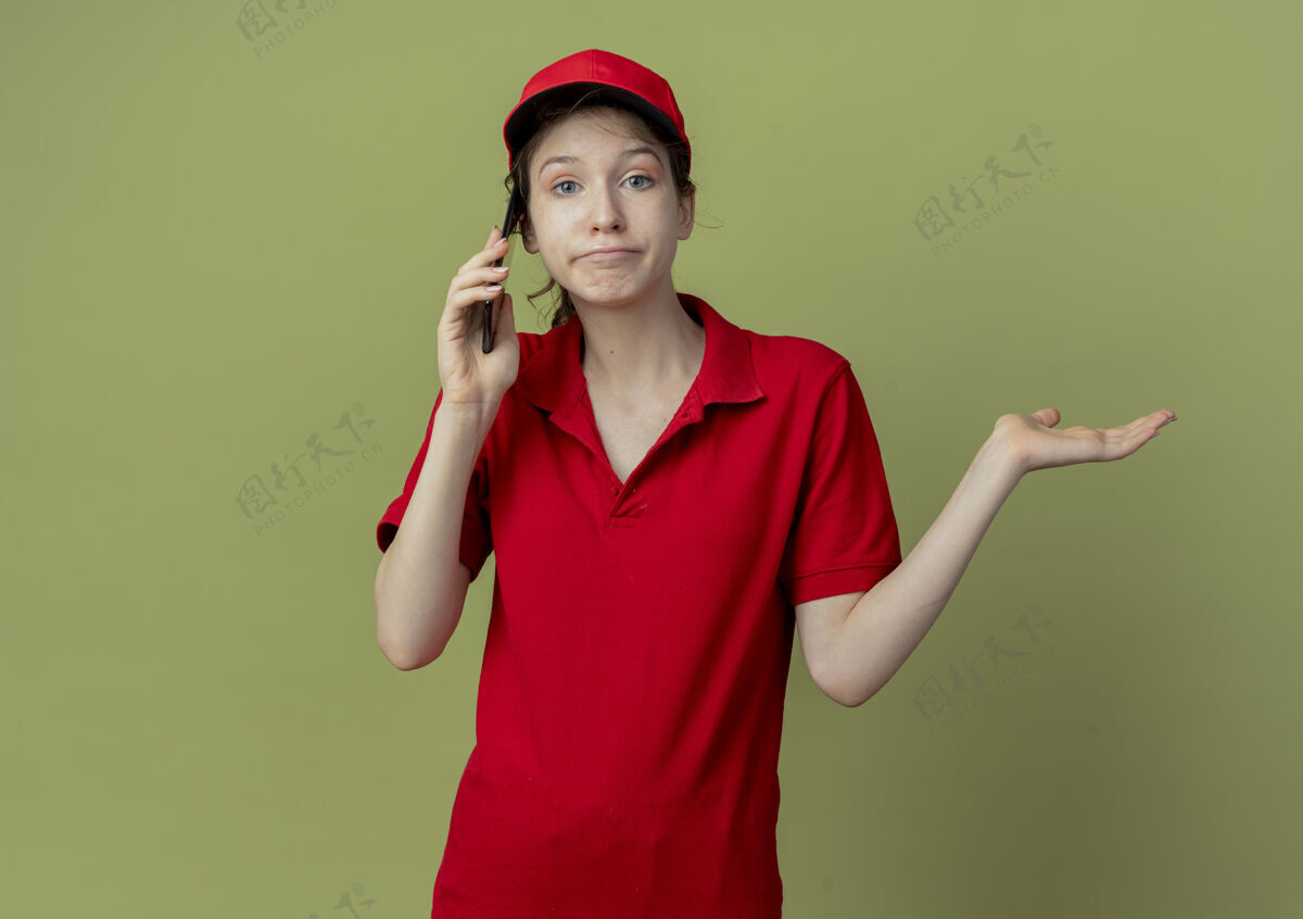 制服困惑的年轻漂亮的送货女孩 穿着红色制服 戴着帽子 一边打电话 一边在橄榄绿的背景上展示着孤立的空手 还有复印空间橄榄绿色交货