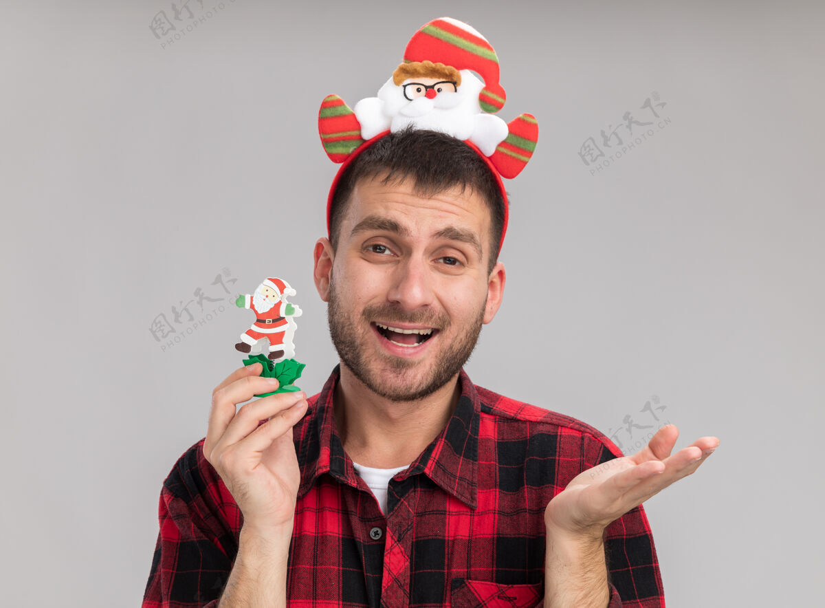 白色印象深刻的年轻白种人戴着圣诞老人头带拿着雪人圣诞玩具看着相机显示空手孤立在白色背景显示人印象
