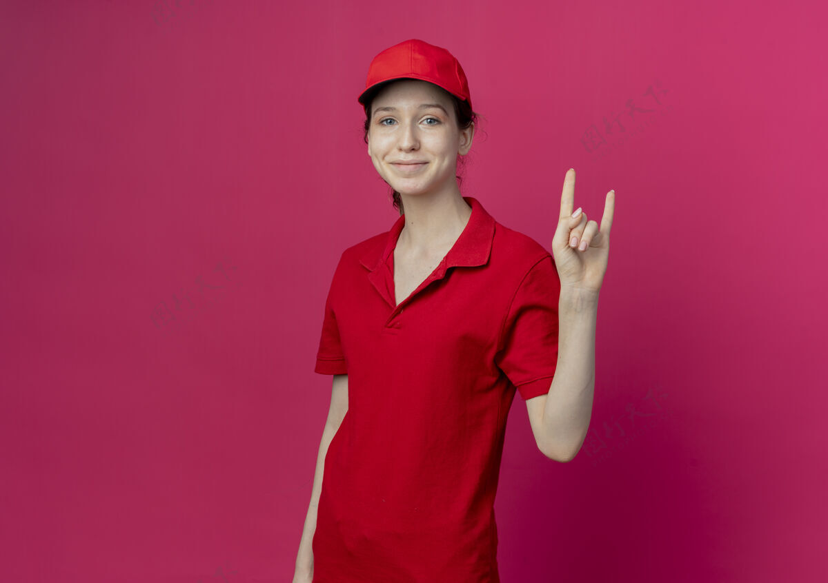 请令人高兴的年轻漂亮的送货女孩穿着红色制服和帽子做岩石标志在深红色的背景与复制空间隔离交货年轻标志