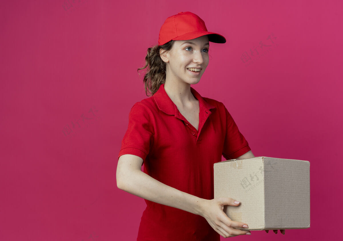 直面带微笑的年轻漂亮的送货女孩穿着红色制服 戴着帽子 看起来笔直地拿着一个纸箱 在深红色的背景上 有复印空间盒子微笑纸箱