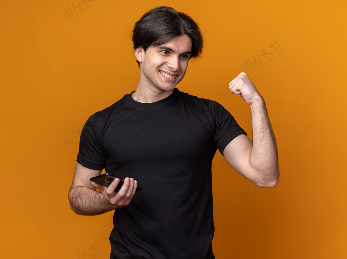 人高兴的年轻帅哥穿着黑色t恤拿着电话 在橙色的墙上显示是的手势孤立市民电话穿着