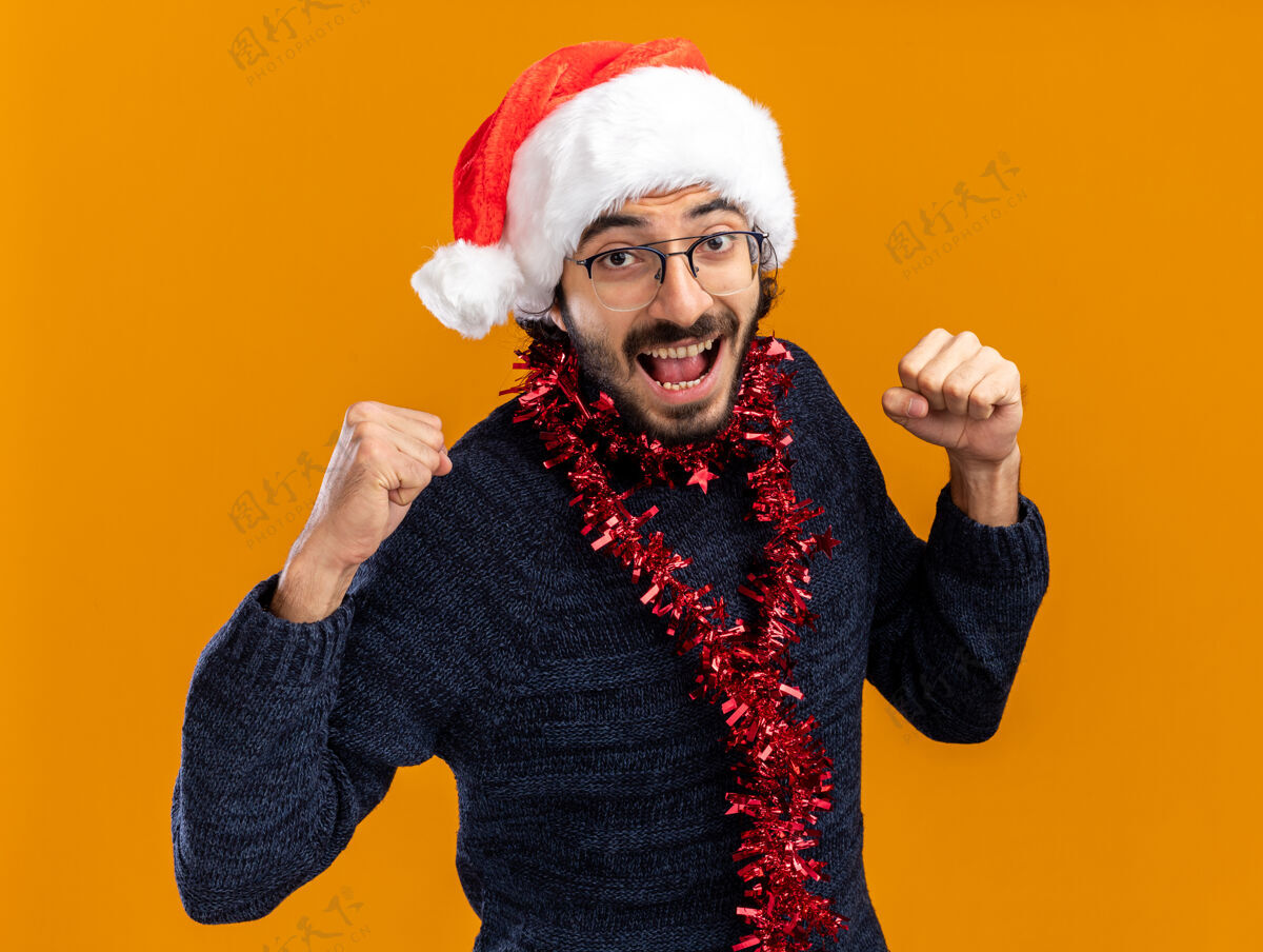 花环兴奋的年轻帅哥戴着圣诞帽 脖子上戴着花环 在橙色背景上显示出“是”的手势橙色年轻圣诞快乐