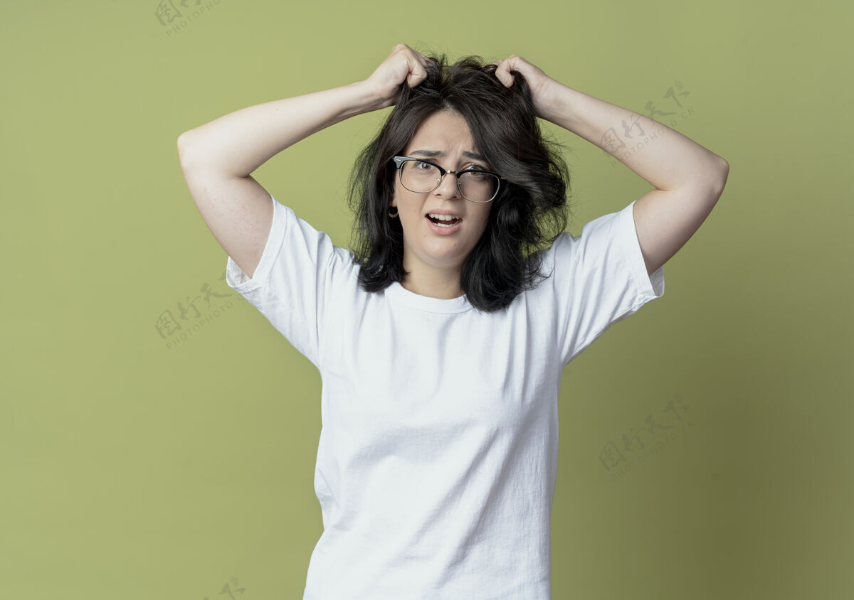 头发印象深刻的年轻漂亮的白人女孩戴眼镜拉她的头发孤立在橄榄绿的背景绿色漂亮眼镜