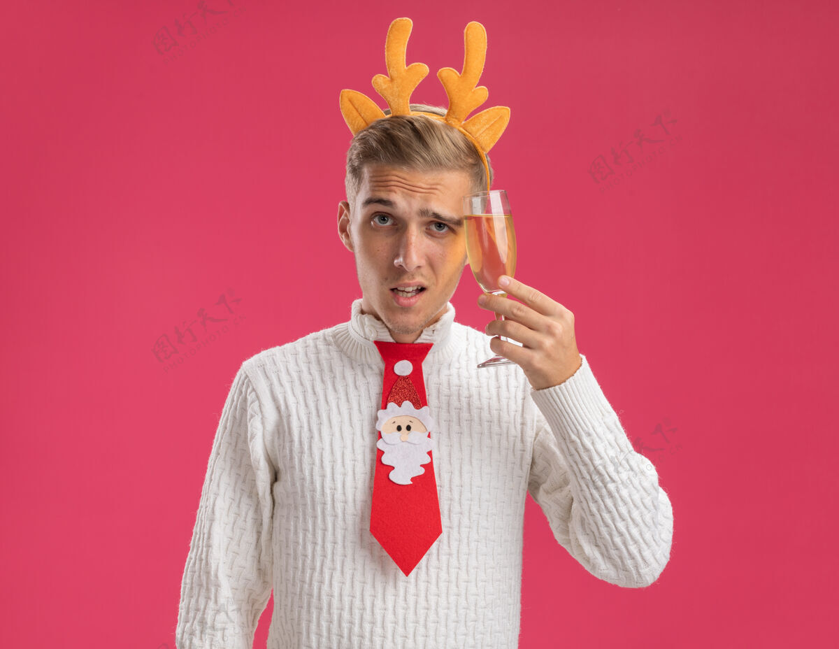 鹿角可疑的年轻帅哥戴着驯鹿鹿角头带 打着圣诞老人的领带 拿着一杯香槟 用它抚摸着头 看着隔离在粉色背景上的相机粉红圣诞快乐领带