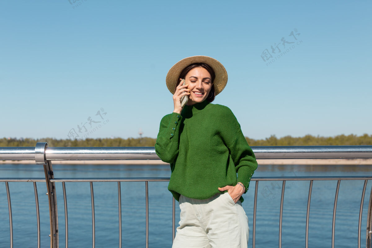 放松穿着绿色休闲毛衣 戴着帽子的时髦女人在户外的桥上 在温暖阳光下的夏日用手机聊天欢乐休闲人