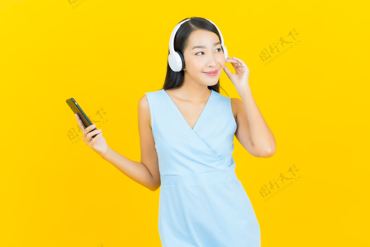 享受画像美丽的亚洲年轻女子戴着耳机和智能手机在黄色的墙上听音乐休闲单人女士