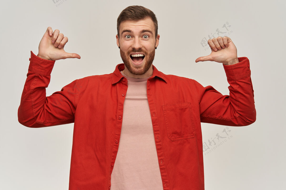 肖像一个身穿红衫 满脸胡须 兴高采烈的年轻人站着 用两只大拇指指着自己 双手放在白墙上发呆意外嘴
