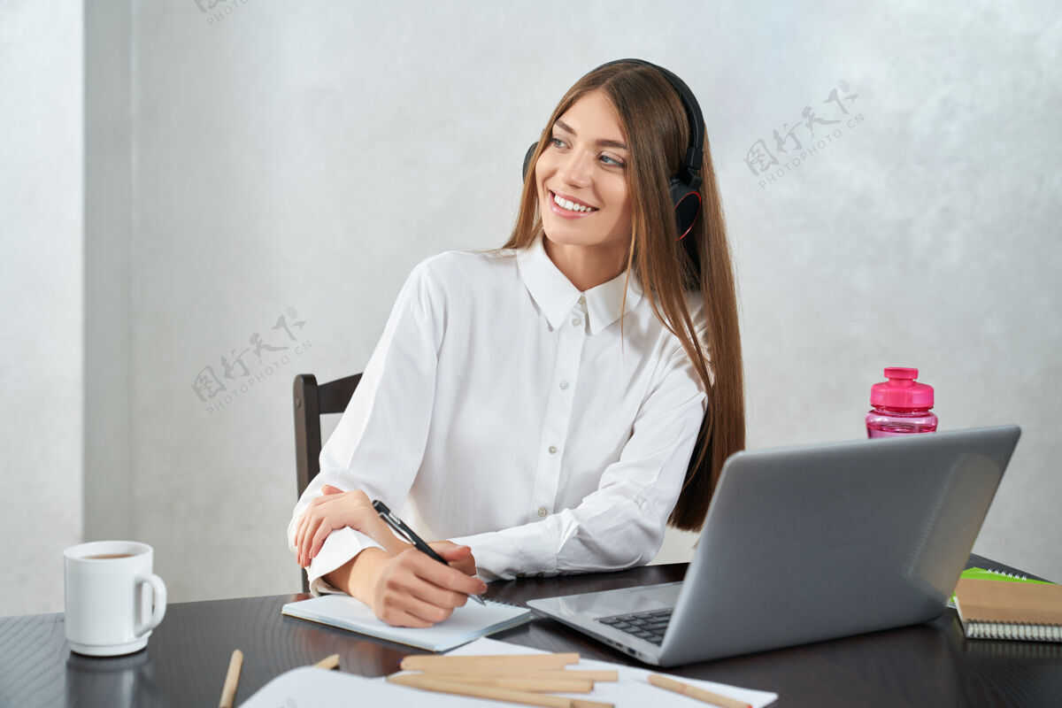 桌子戴着耳机在笔记本电脑上学习的积极的女人学生年轻耳机