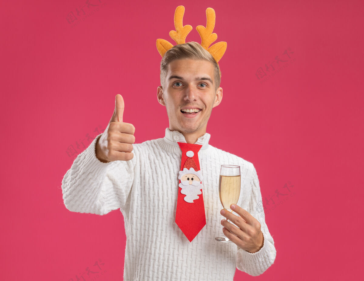 领带快乐的年轻帅哥戴着驯鹿鹿角头带 打着圣诞老人的领带 手里拿着一杯香槟 看着相机 在粉色背景上孤立地竖起大拇指玻璃粉红圣诞老人