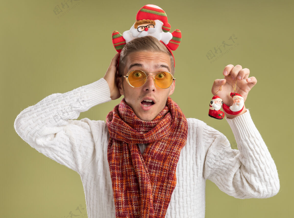 年轻印象深刻的年轻帅哥戴着圣诞老人的头带和围巾拿着圣诞老人的圣诞饰品看着相机保持手在头后橄榄绿的背景隔离装饰家伙举行