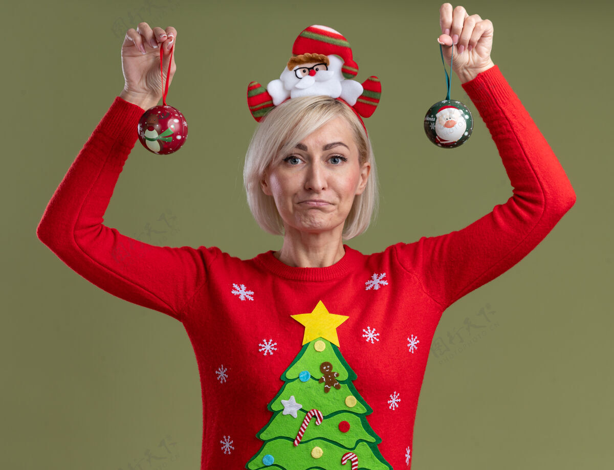头带笨手笨脚的中年金发女人戴着圣诞老人的头带和圣诞毛衣举起圣诞饰品看着橄榄绿背景上孤立的相机无知加薪金发