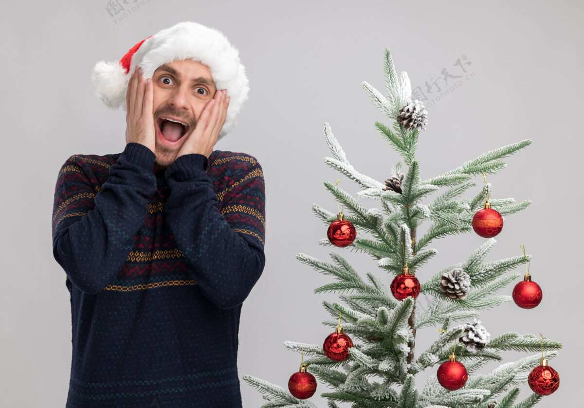 男人兴奋的白人年轻人戴着圣诞帽站在圣诞树旁 手放在脸上看着隔离在白色背景下的摄像机白色帽子手