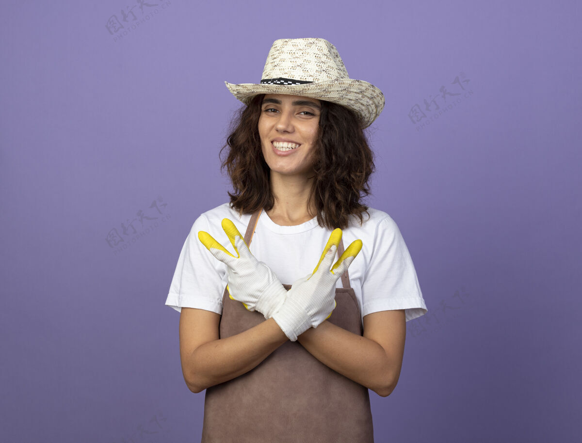 成人身着制服的年轻女园丁微笑着戴着园艺帽和手套 双手交叉 表现出和平的姿态人园艺年轻