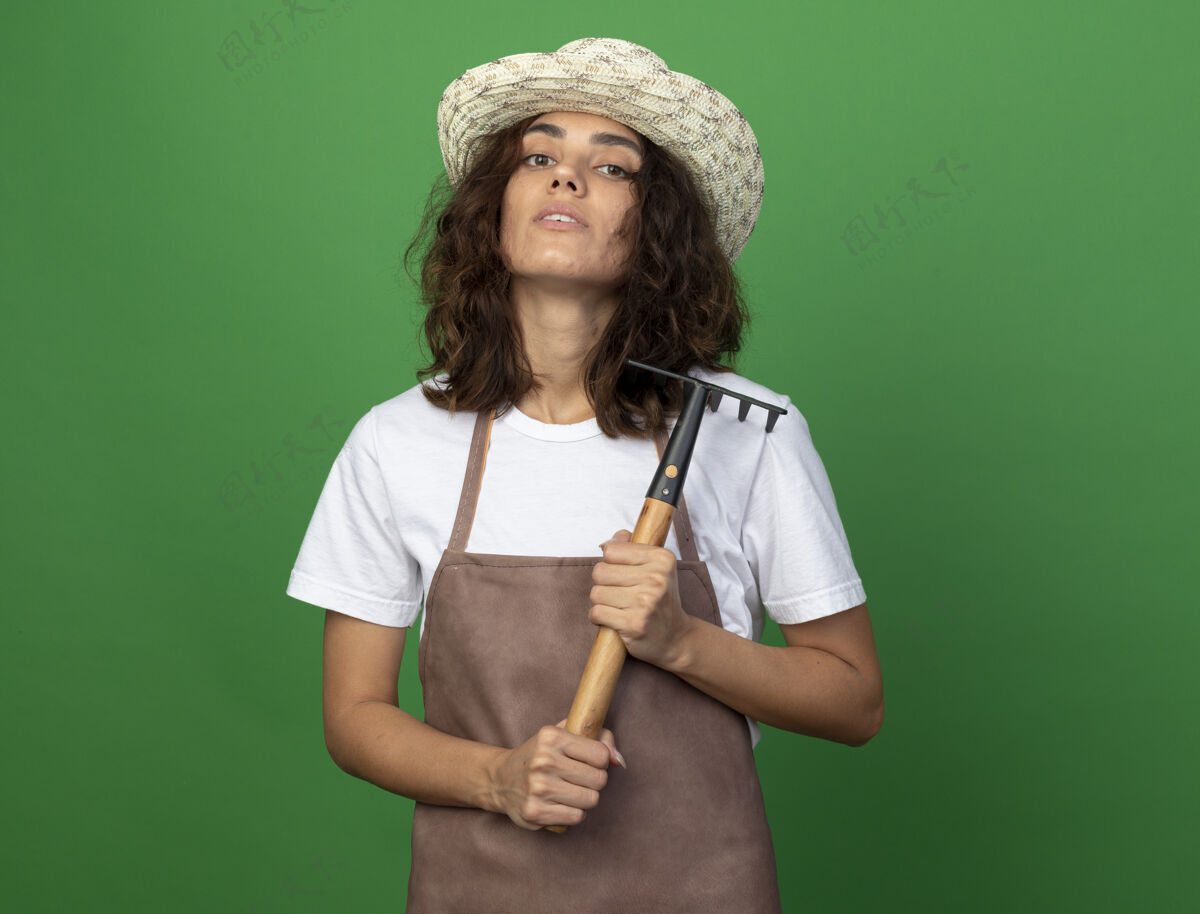 举行自信的年轻女园丁穿着制服戴着园艺帽拿着耙子人帽子女人