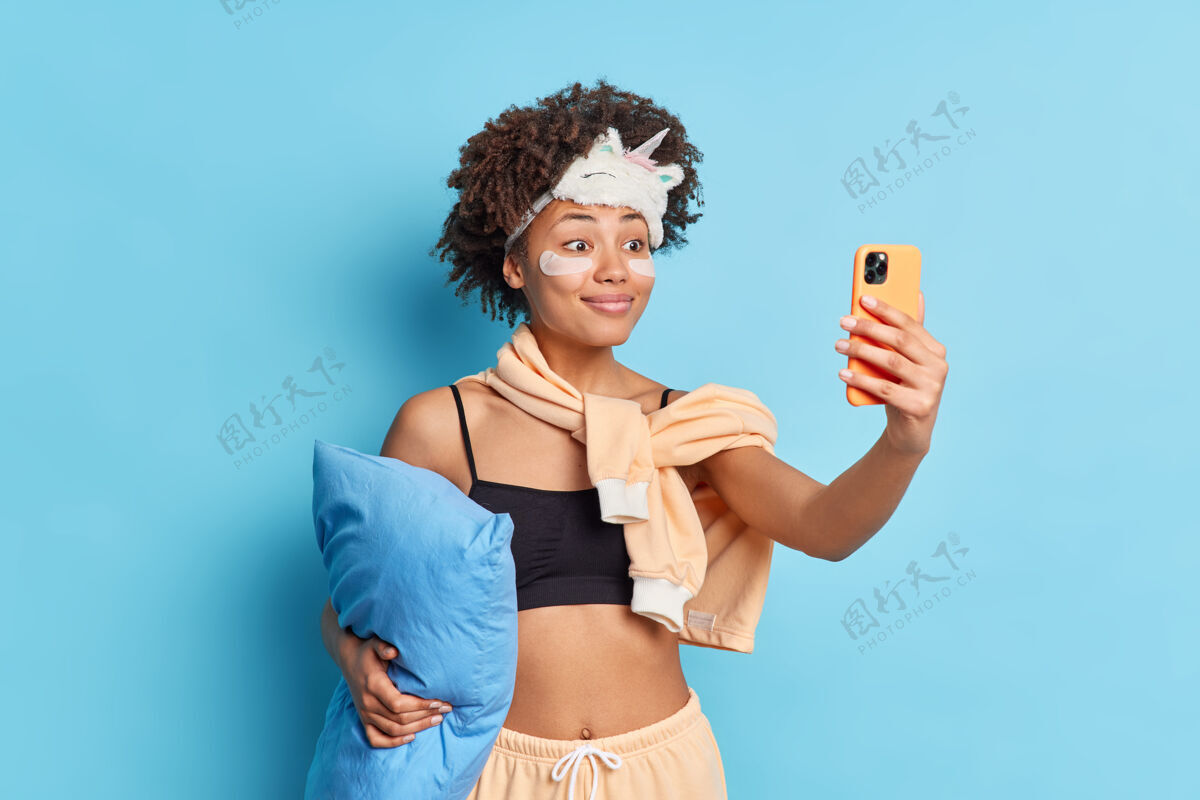 成人穿着睡衣 面带微笑的美籍非洲妇女 戴着睡眠面罩 抱着枕头 通过手机自拍 贴着胶原蛋白贴片请女性蒙眼