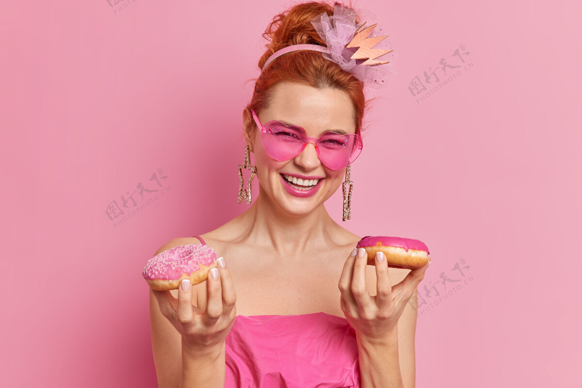 甜甜圈高兴的红发女人表达积极的心情捧着两个开胃的甜甜圈开心地笑着心情好女孩高兴粉色