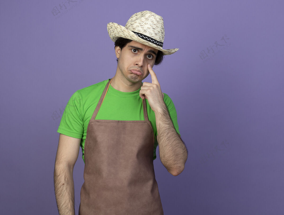 紫色悲伤的年轻男园丁穿着制服戴着园艺帽把手指放在眼睛上手指眼睛帽子