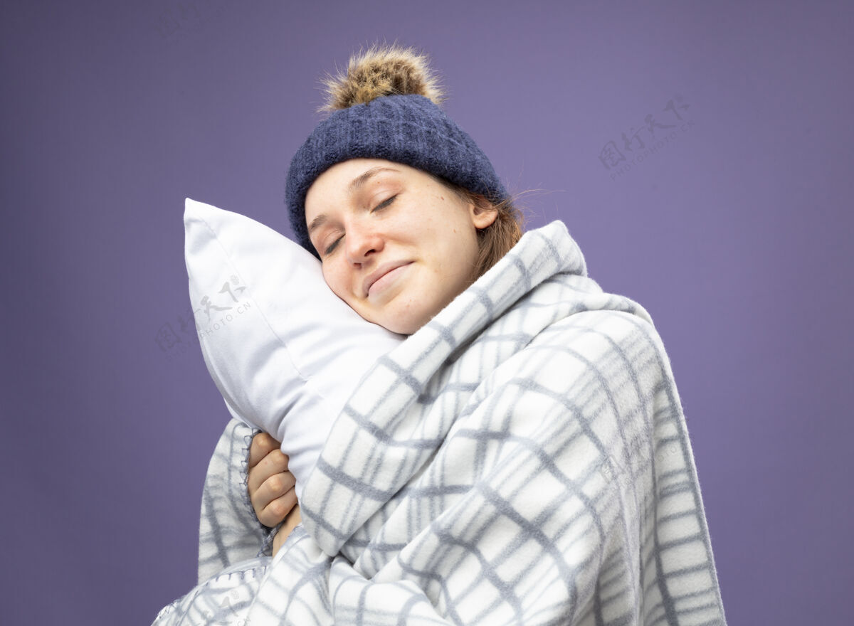 年轻高兴的年轻病女孩闭着眼睛穿着白色长袍和冬季帽子围巾包裹在格子抱枕眼睛拥抱女人