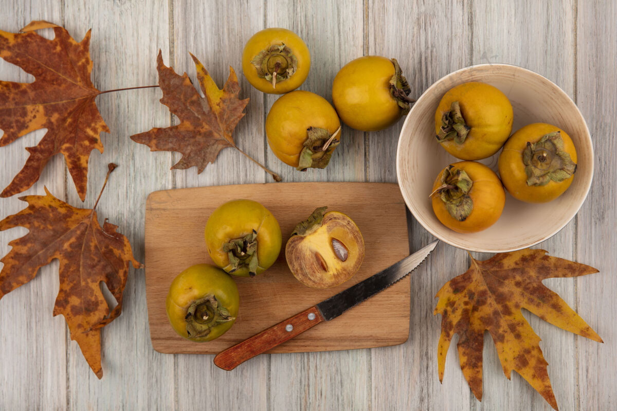 橘子黄色柿子水果的顶视图在一个碗上 叶子和柿子水果放在一个木制的厨房板上 刀放在灰色的木桌上刀木头厨房