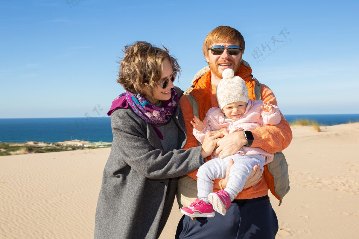 休闲快乐的父母抱着可爱的小女儿站在沙滩上 在海上欢呼爱为人父母