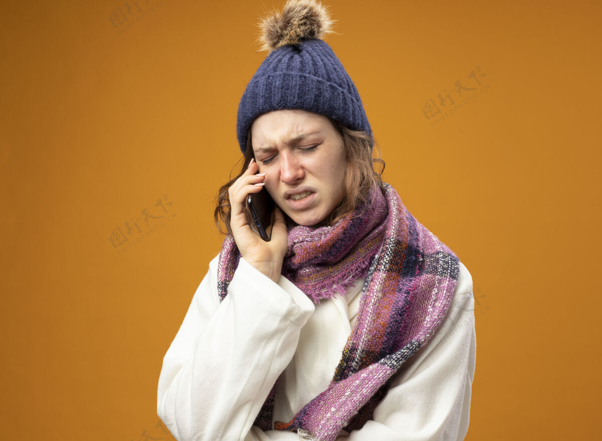 不愉快戴着白色长袍 戴着围巾的冬帽 闭着眼睛的未被释放的年轻病女孩在橙色的墙上讲电话电话长袍女孩
