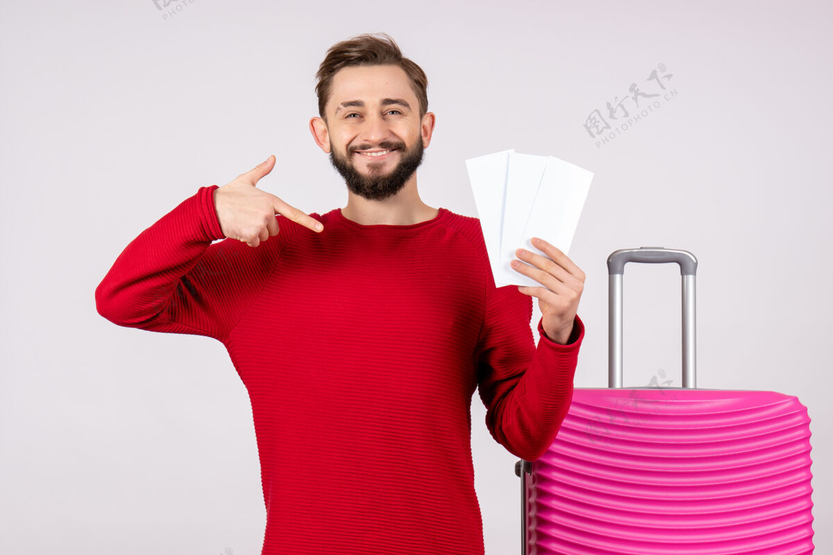 微笑正面图：年轻男性 带着粉色包 手持飞机票 在白墙上航行 飞行旅行 度假 情感照片 游客机票成人游客
