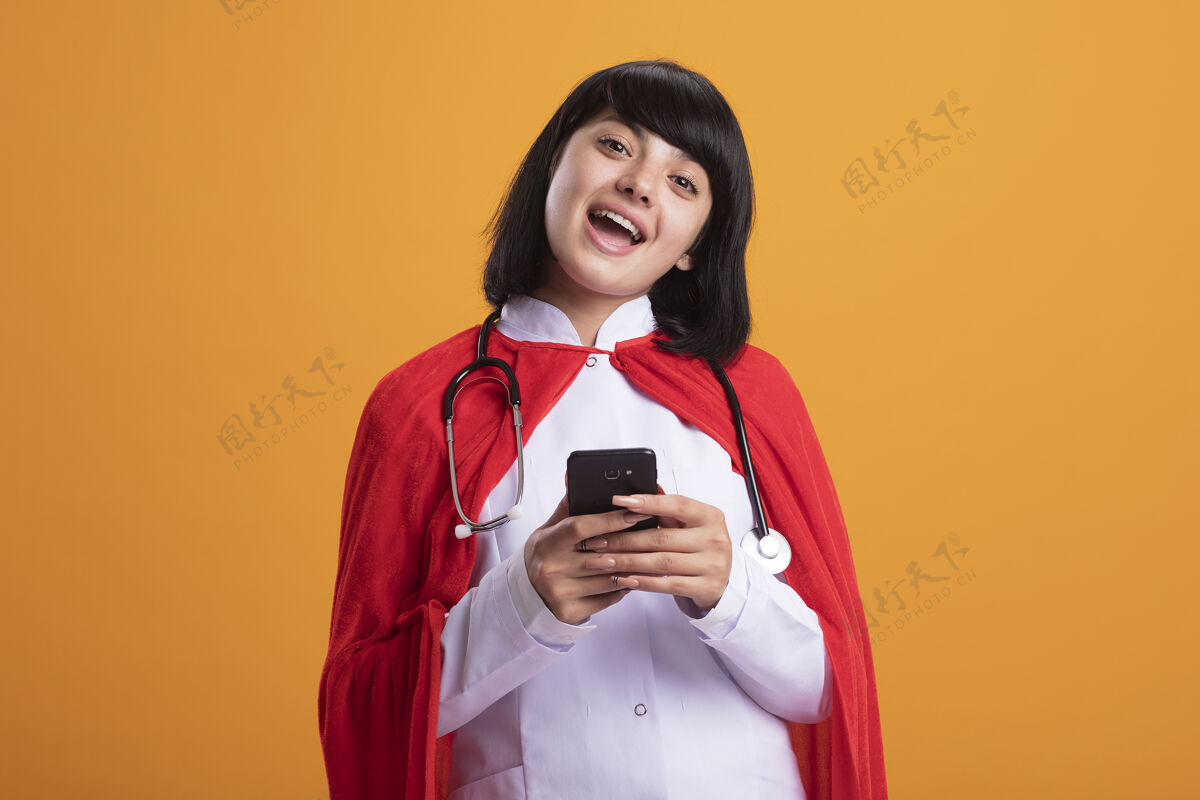 年轻带着微笑的年轻超级英雄女孩戴着听诊器 穿着医用长袍和斗篷 把电话隔离在橙色的墙上长袍医疗穿