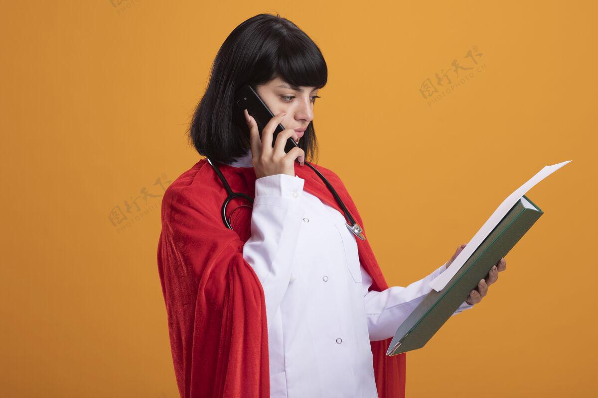 年轻想着穿着医用长袍和斗篷的听诊器的年轻超级英雄女孩在打电话 看着孤立在橙色墙上的手上的纸思考斗篷女孩