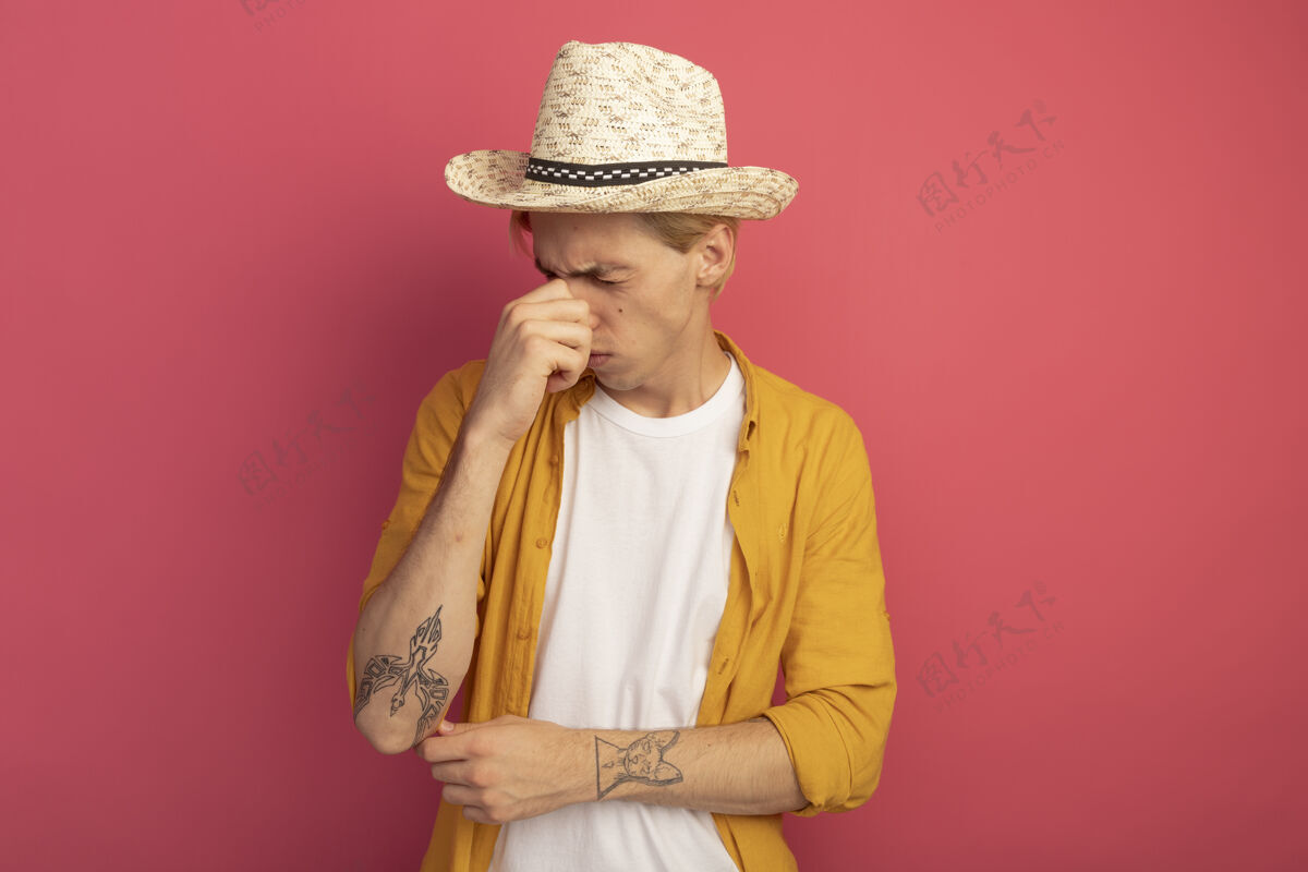 黄色戴着黄色t恤和帽子 闭着眼睛的年轻金发男人抓住了粉红色的鼻子粉色帽子戴着