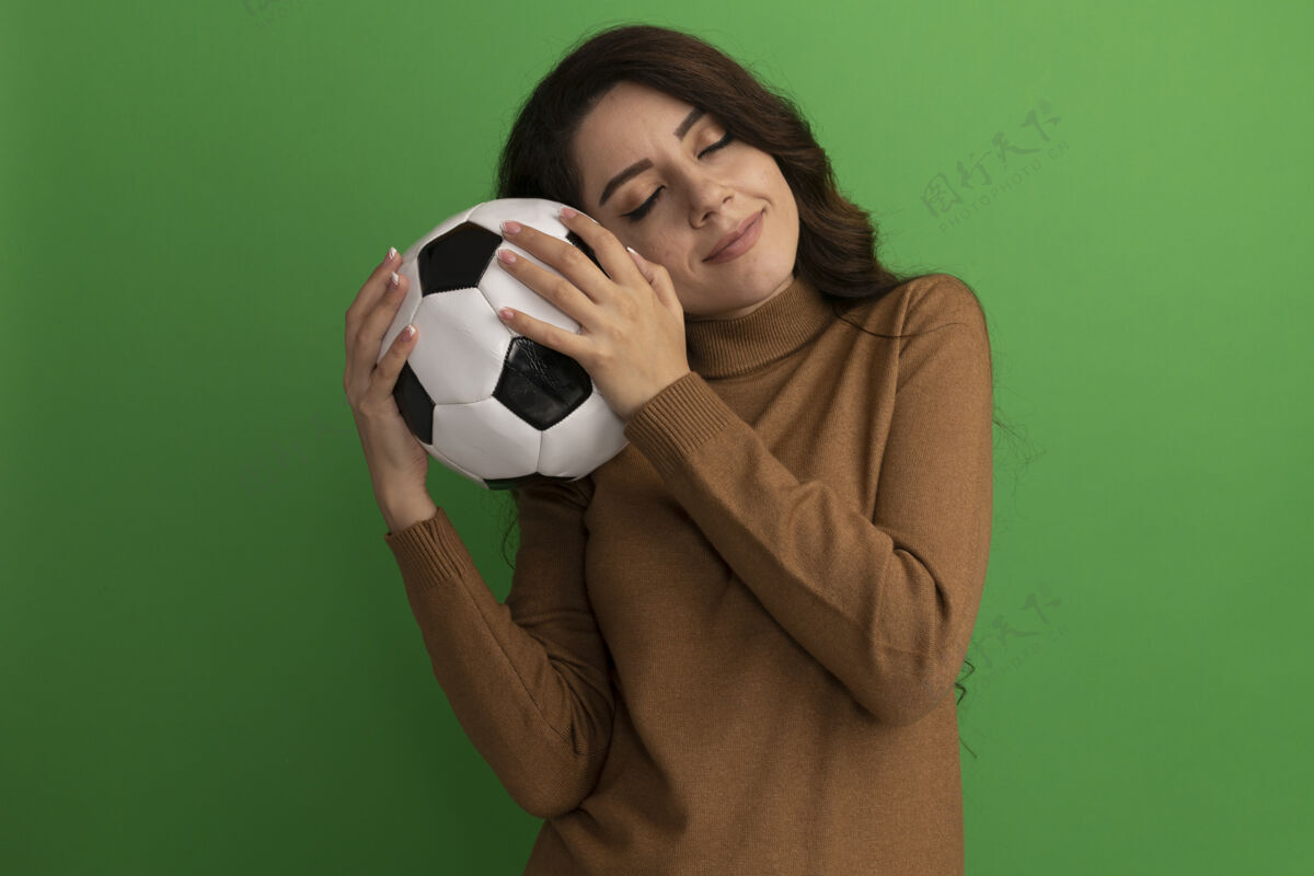 女孩高兴地闭上眼睛年轻漂亮的女孩抱着球在脸颊上隔离在绿色的墙壁上闭上脸颊球