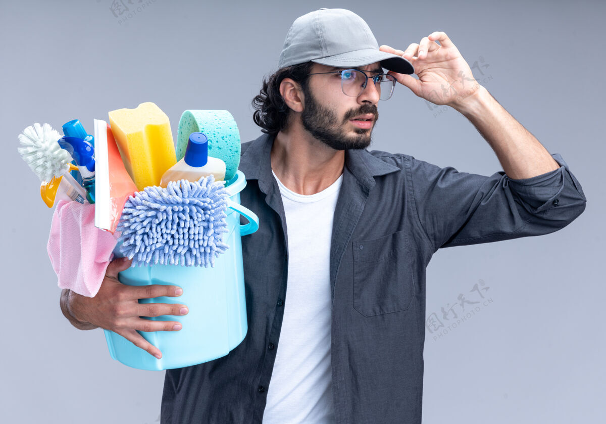 持有迷茫的年轻帅哥 穿着t恤 戴着帽子 手里拿着一桶清洁工具 手里拿着帽子 隔离在白色的墙上帽子桶工具