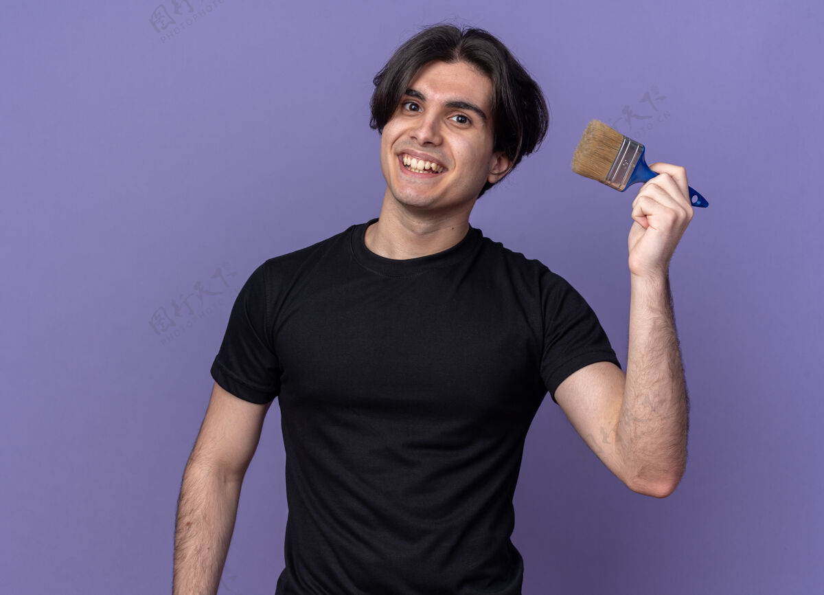抱着微笑的年轻帅哥穿着黑色t恤拿着油漆刷孤立在紫色的墙上穿微笑小伙子