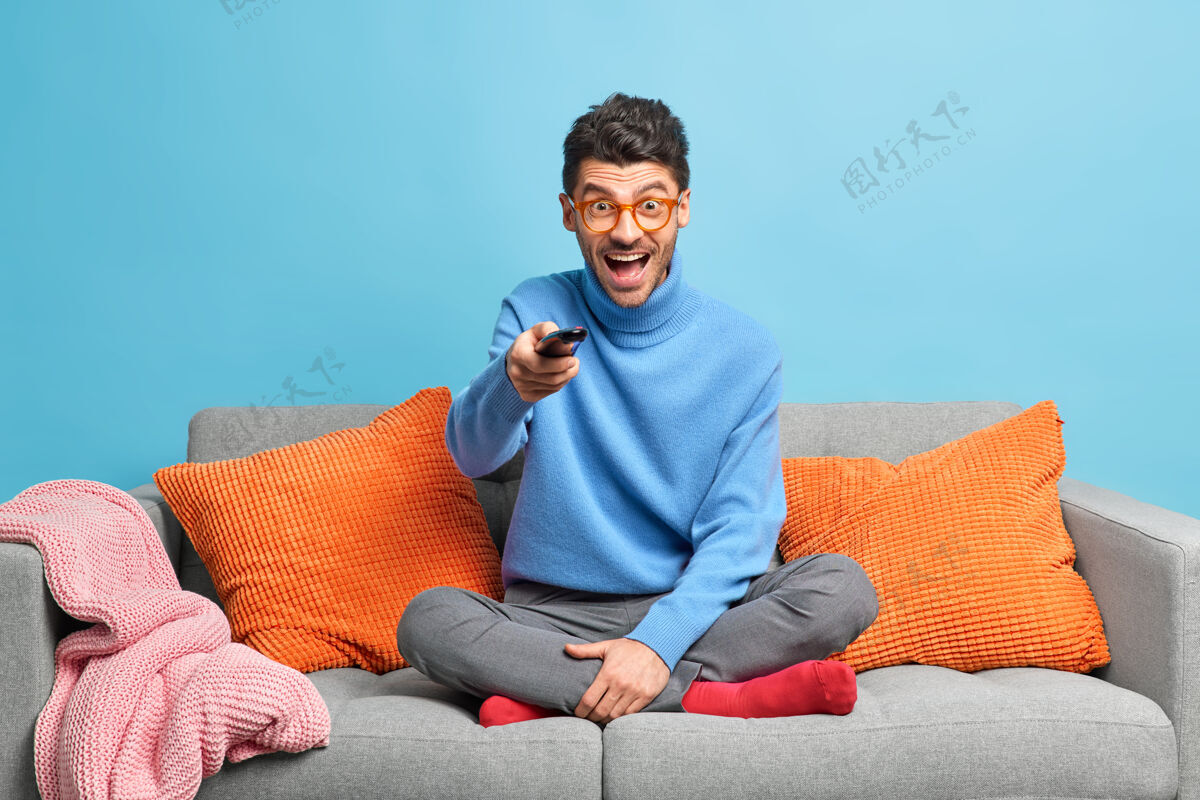 遥控器人们休闲消遣的概念喜出望外的未刮胡子的成年男子坐在沙发上摆莲花姿势 手持遥控器 在电视上看搞笑节目一个控制手表