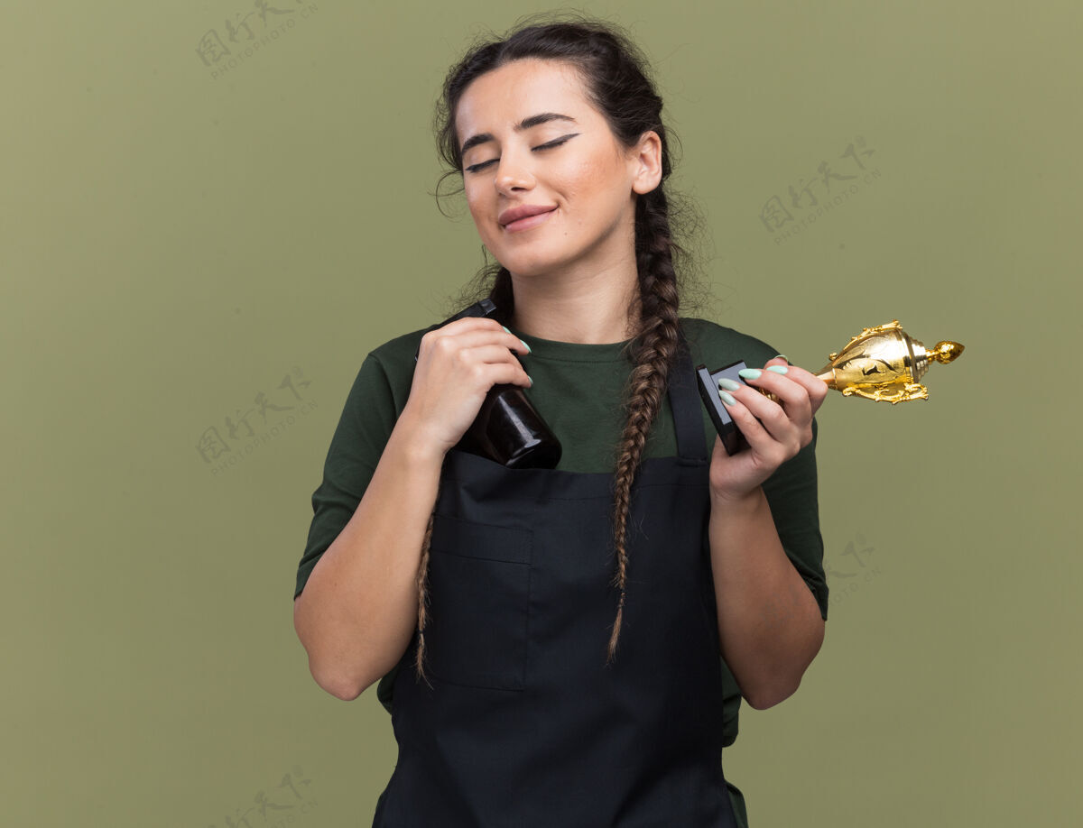 橄榄色高兴地闭上眼睛年轻的女理发师在统一举行优胜者杯与理发师隔离在橄榄绿的墙上理发师年轻手持